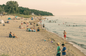 Beach goers dot the sandy shoreline, with trees in the background and lake on right side